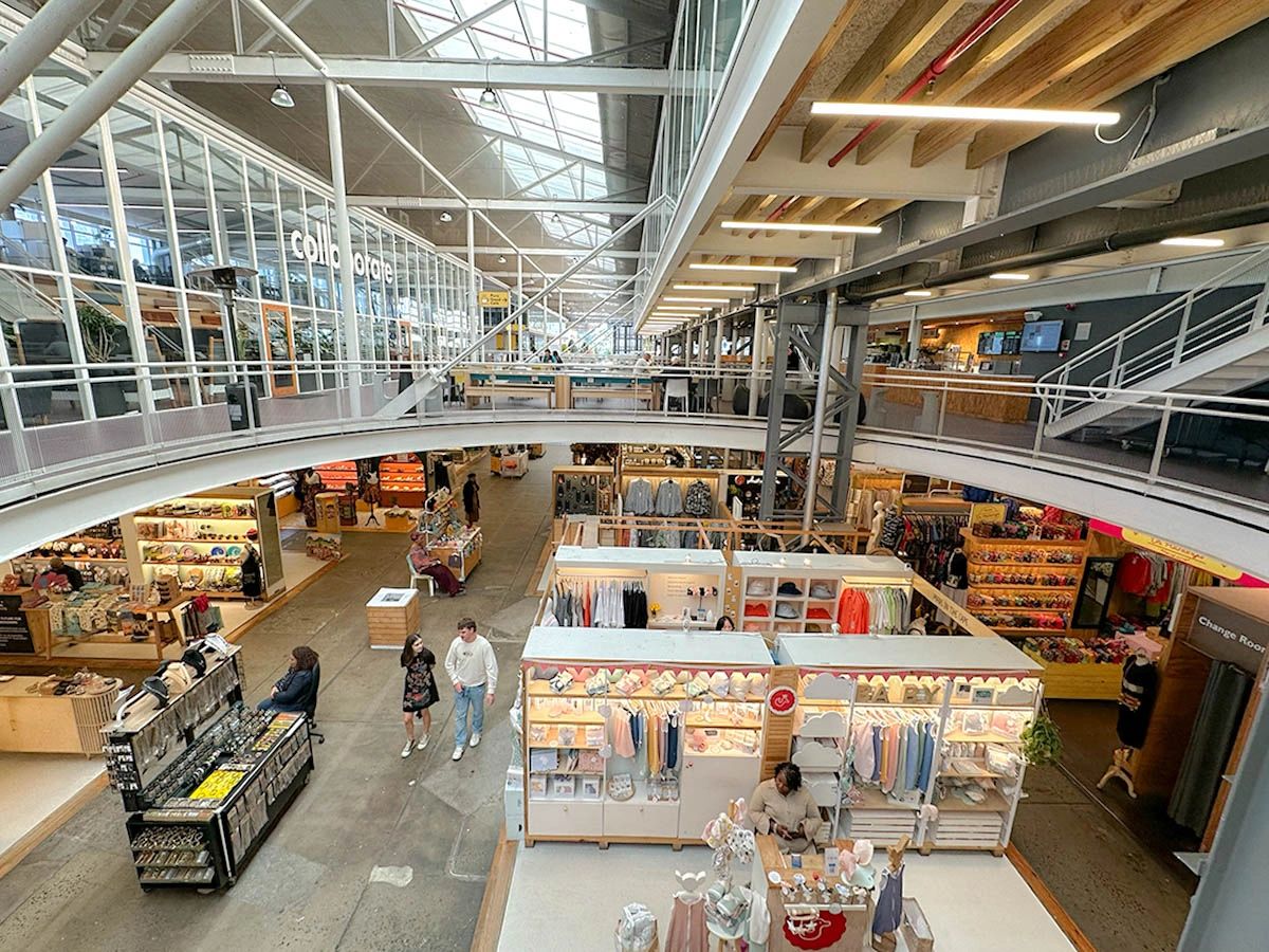Interior view of the V&A Waterfront’s Watershed Market in Cape Town, showcasing its modern industrial design with exposed steel beams, skylights, and a mezzanine level. Small retail stalls offer local crafts, clothing, jewelry, and handmade goods. Shoppers stroll between brightly lit booths in the spacious open-plan layout. I Marketplace interiorWatershed at V&A Waterfront interior with local artisan vendors and modern industrial architecture in Cape Town