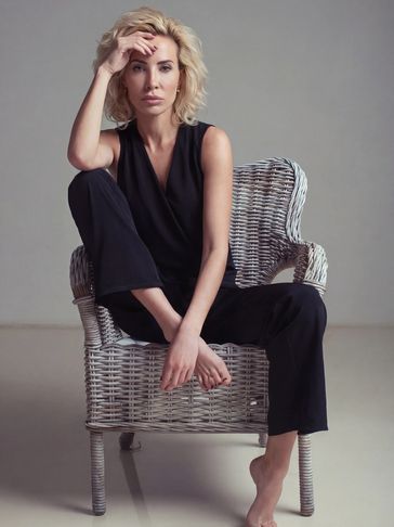 A blonde woman in black outfit sitting thoughtfully on a wicker chair.