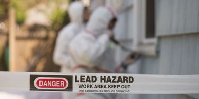 Danger tape indicating lead hazard with workers in protective suits in the background.