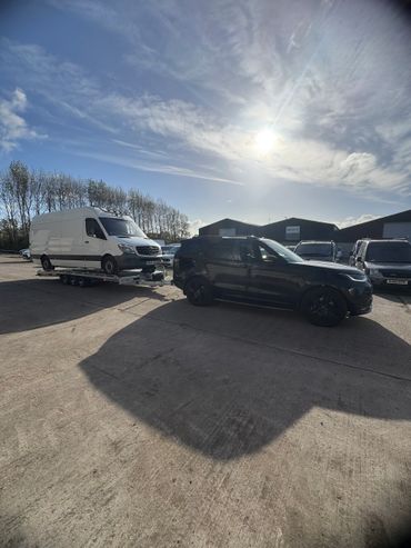 Black SUV towing a white van on a trailer in a parking lot under a bright sky.