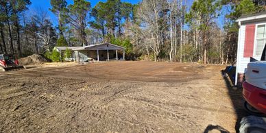 Cleared dirt lot with trees and a carport in the background on a sunny day.