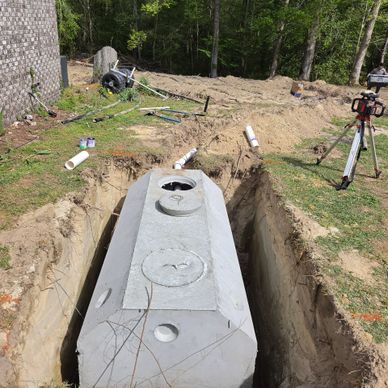 Concrete septic tank being installed in a dug trench beside a house.