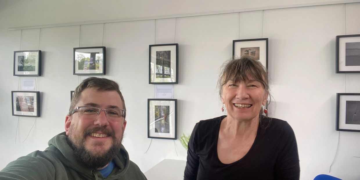 Two people smiling in a room with framed photos on the wall.