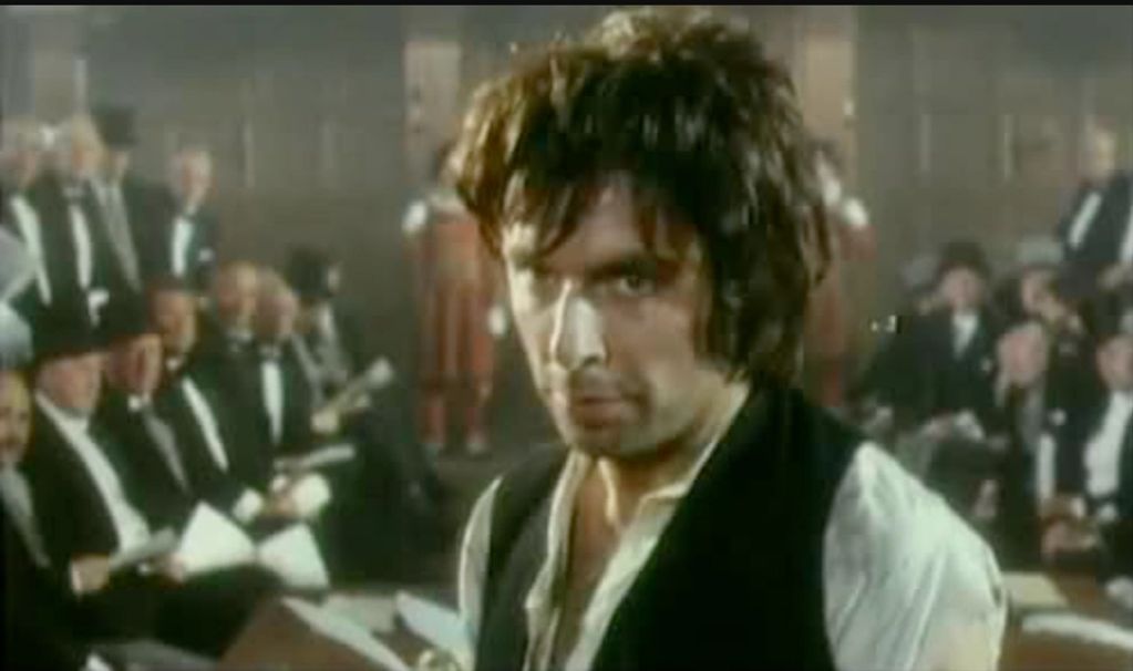 A man in old-fashioned clothes looks intensely forward in a formal gathering.