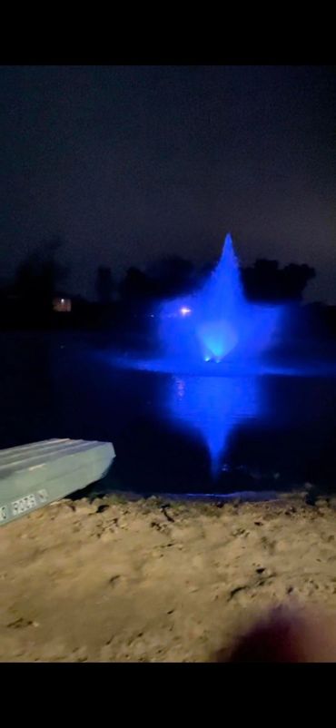 Blue-lit fountain glowing on a lake at night with a small boat on the shore.