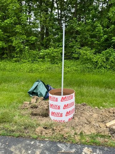 A cylindrical concrete form with a white pole in a grassy area near a driveway.