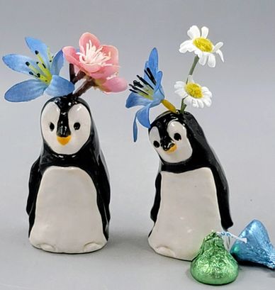 Two ceramic penguin figurines with flowers on their heads and candy beside them.