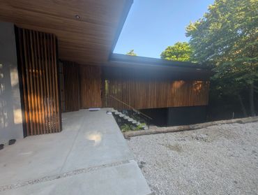 Modern house entrance with wood paneling and outdoor stairs surrounded by trees.
