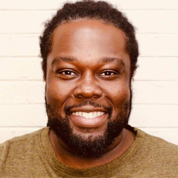 Smiling man with a beard against a light brick wall.