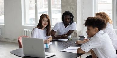 Group of students gathered around a table with a laptop, practicing conversation and language skills