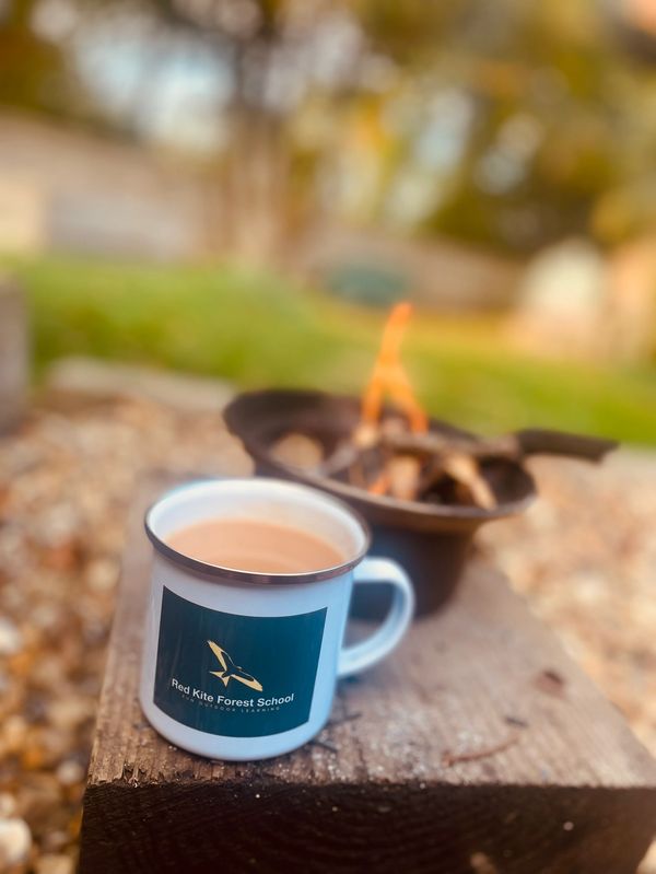 A mug of hot drink beside a small outdoor fire on a wooden block.