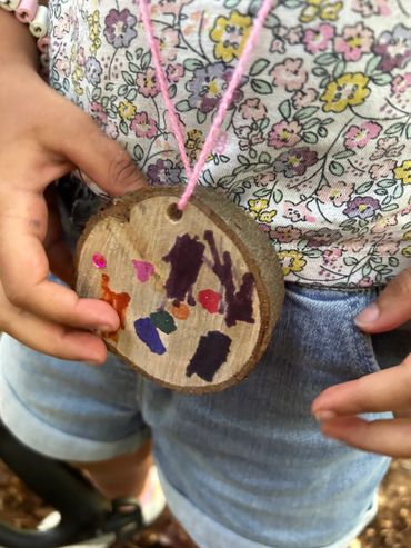 Child holding a handmade wooden pendant with colorful drawings and a pink string.
