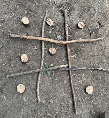 Tic-tac-toe game made from sticks and wood pieces on dirt.