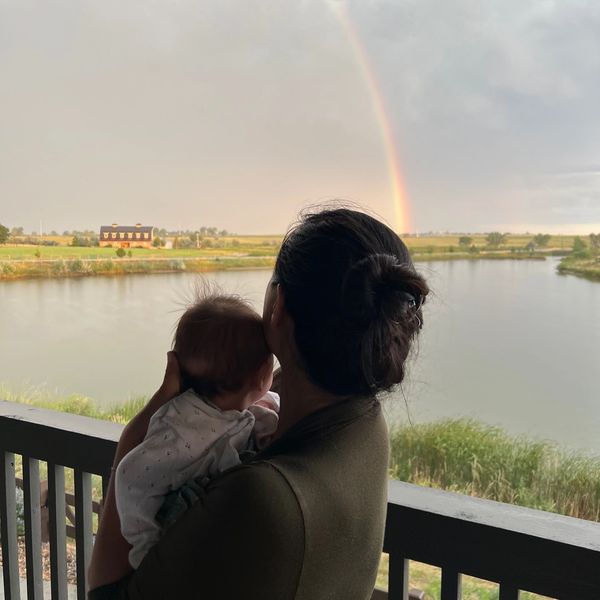 A woman holding a baby, gazing at a rainbow over a serene lake.