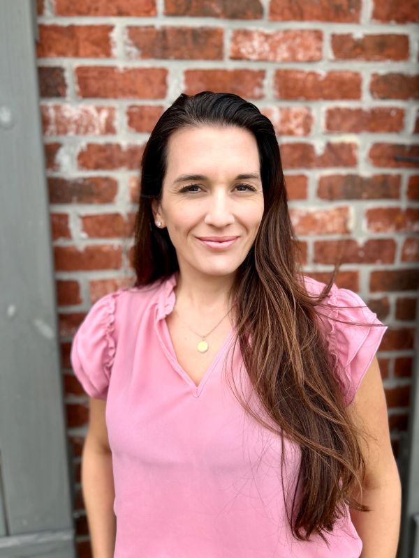 Woman with long brown hair in a pink blouse standing against a brick wall.