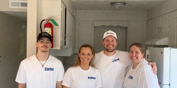 Four people wearing matching white shirts with names pose in a kitchen.