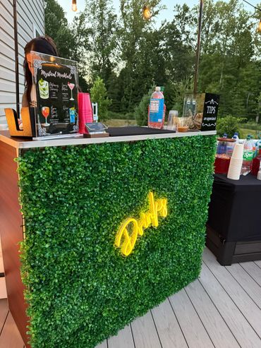 Outdoor drink bar with greenery and neon sign.