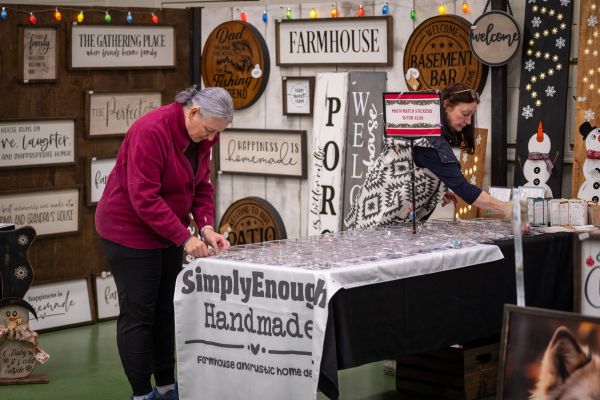 Two women browsing handmade farmhouse decor and stickers at a market stall.