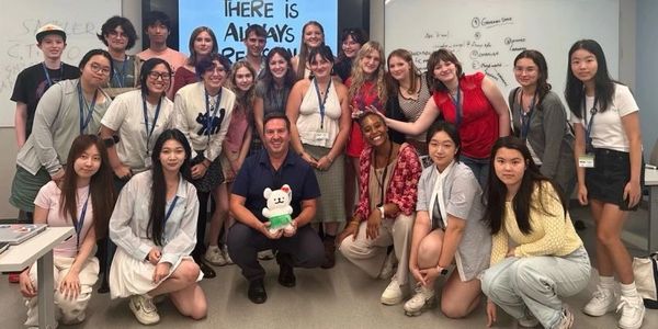 A diverse group of young adults and a man posing together in a classroom.