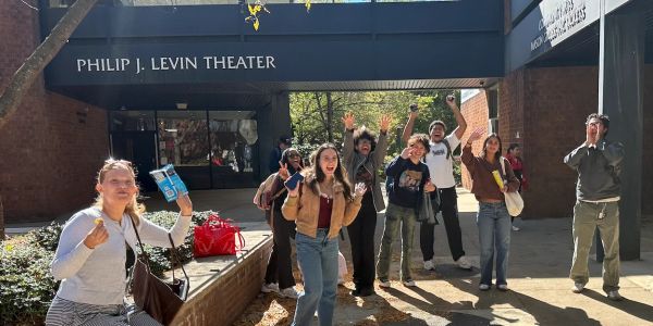 Group of young people cheering outside Philip J. Levin Theater on a sunny day.