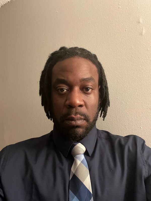Man with dreadlocks wearing a navy shirt and blue patterned tie.