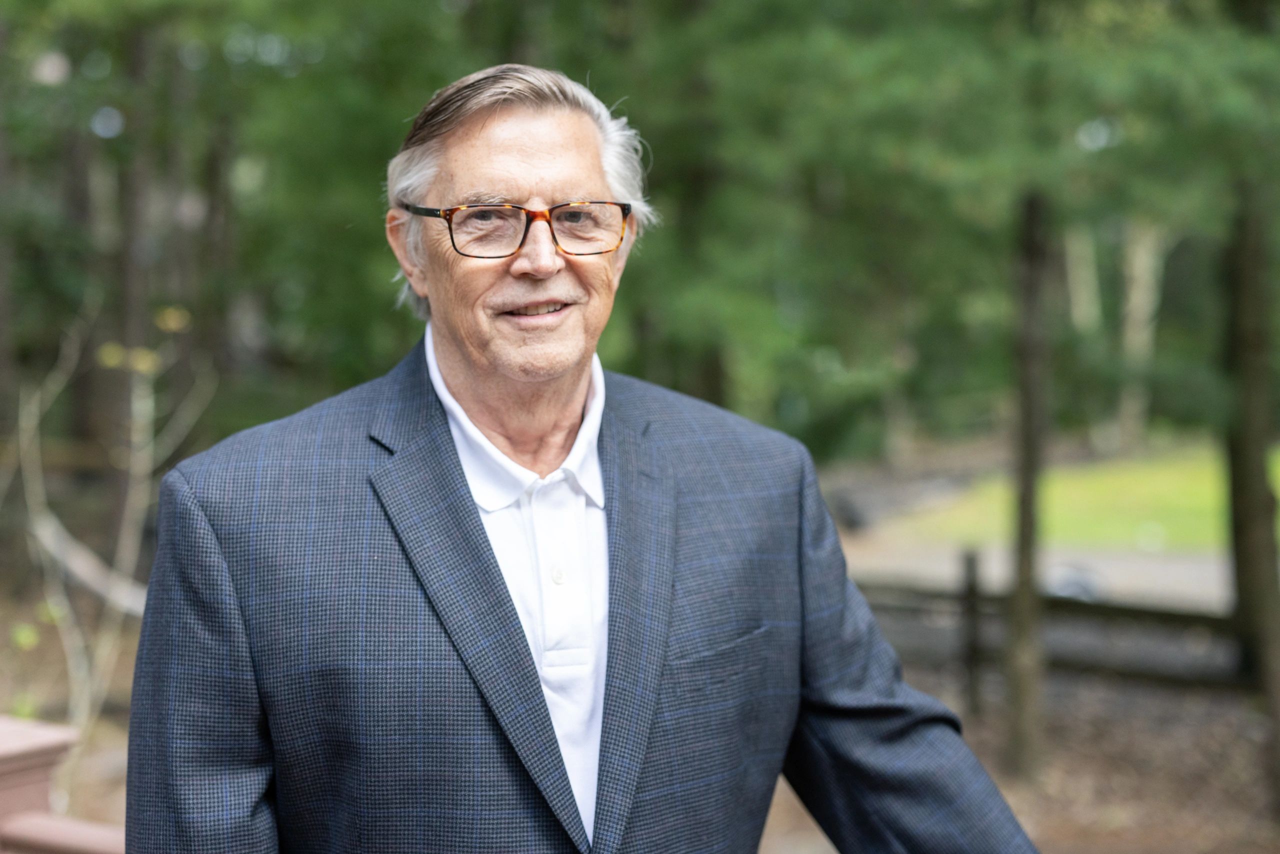 Smiling elderly man in glasses and a checkered blazer outdoors.