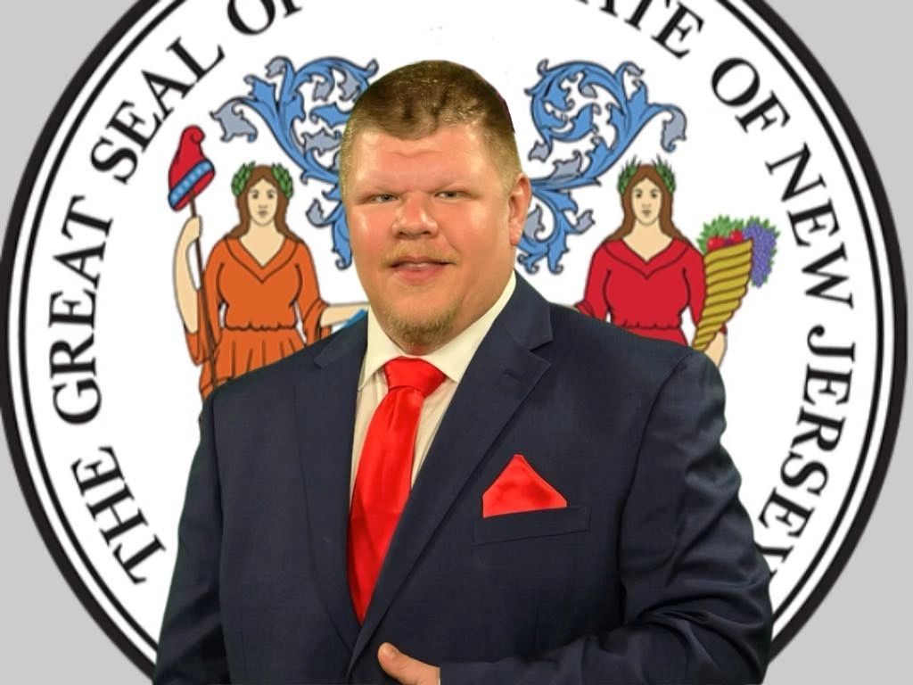 Man in suit with red tie and pocket square in front of New Jersey state seal.