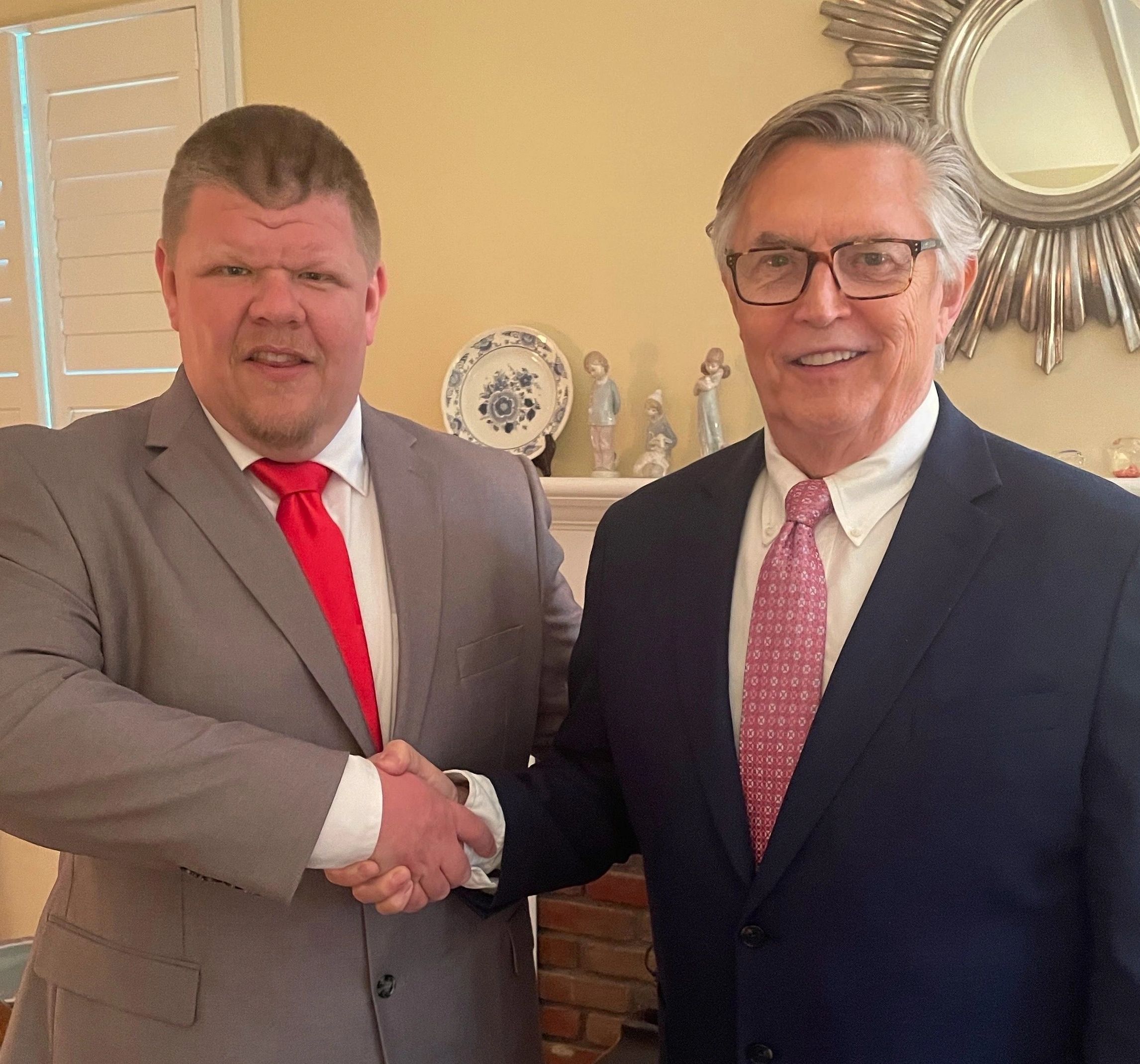 Two men in suits shaking hands indoors, smiling warmly.
