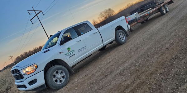 White Ram pickup truck towing a flatbed trailer on a dirt road at sunset.