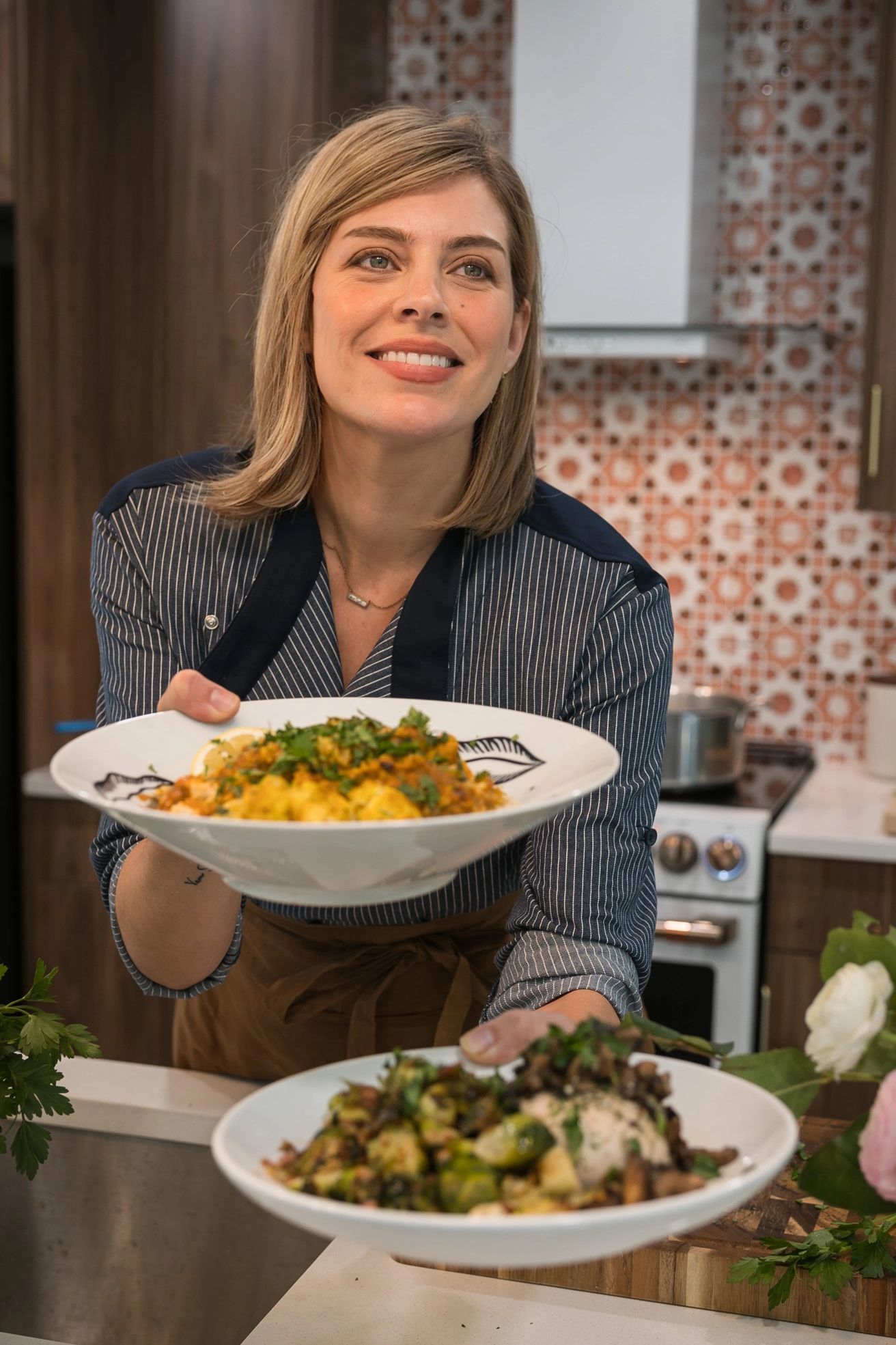 Smiling woman presenting two plates of colorful, fresh food in a cozy kitchen.
