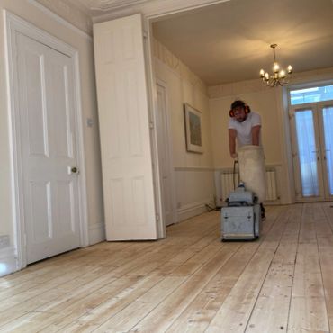 Man sanding wooden floor in a bright room with ear protection.