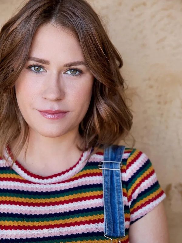 Young woman with wavy brown hair wearing a colorful striped shirt and denim overalls.