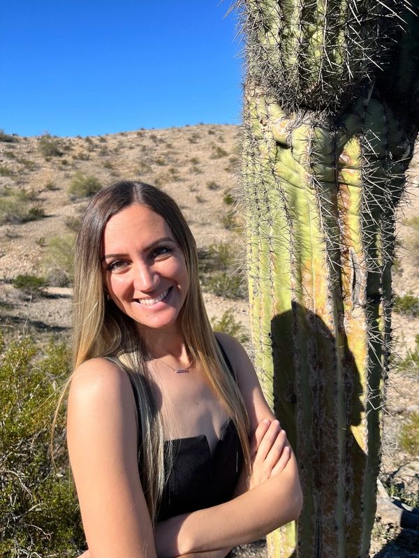 Woman smiling beside a tall cactus in a desert.