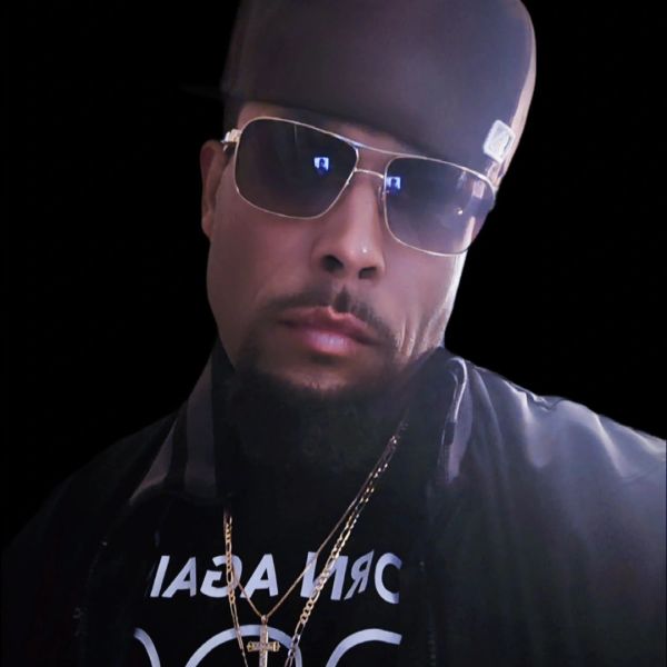 Man wearing sunglasses, a cap, and gold chains against a black background.
