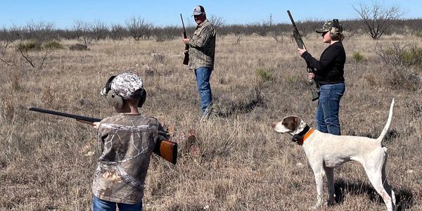 Three people with rifles and a hunting dog in a dry grassy field.