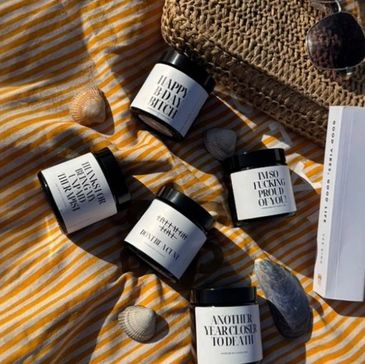 Candles with bold, humorous messages on a striped cloth beside a woven bag and sunglasses.