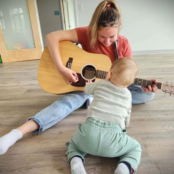A woman plays guitar while a baby reaches out to touch it on a wooden floor.