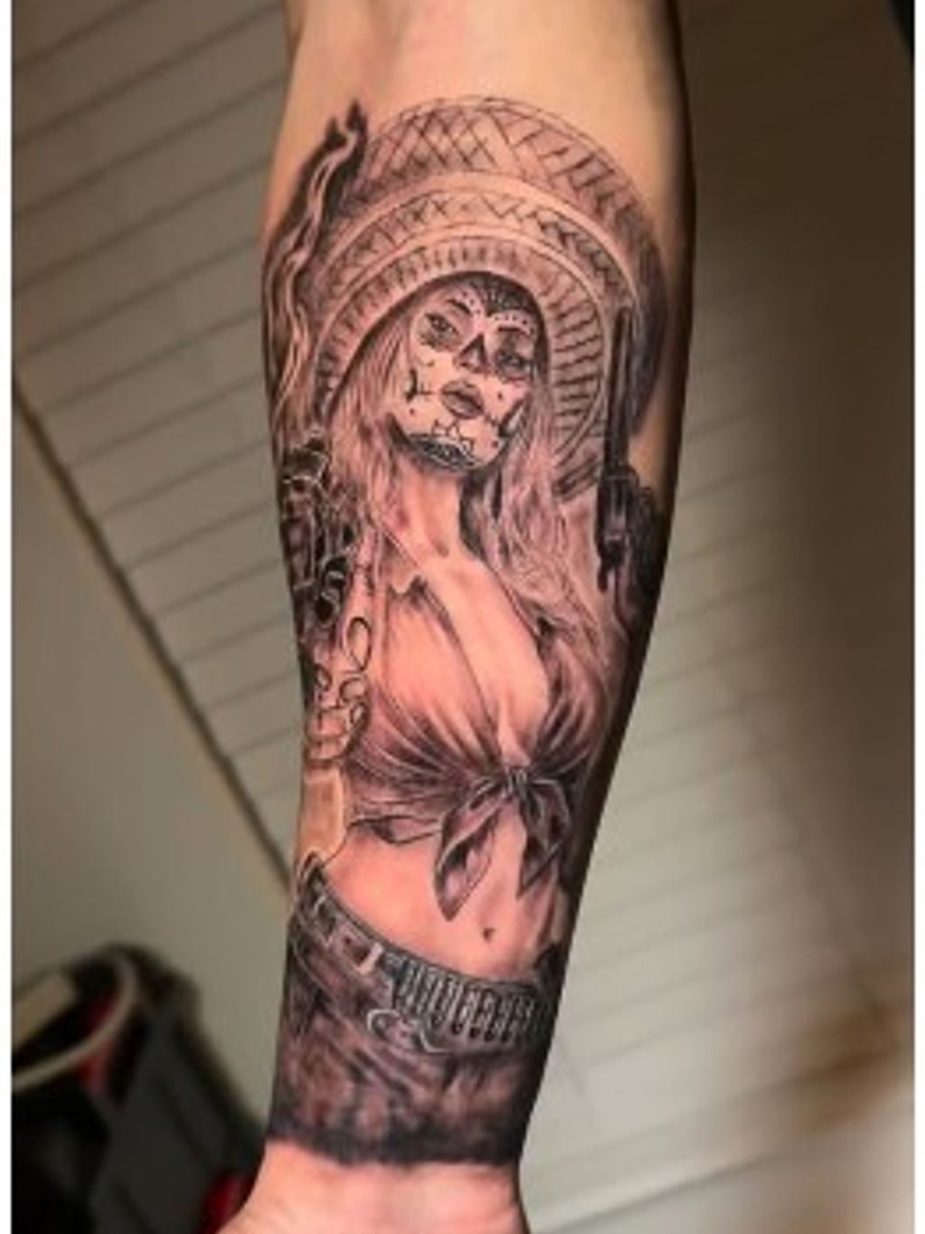 Tattoo of a woman with Day of the Dead face paint holding guns, wearing a tied shirt and sombrero.