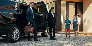 Businessman exiting car greeted by kids and a woman at home.