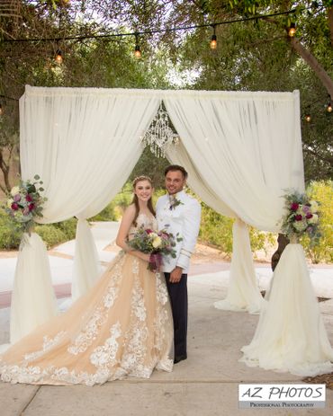 Bride and groom posing under a decorated wedding arch outdoors.
