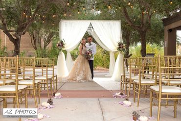 Bride and groom posing under a white draped wedding arch with gold chairs and string lights.