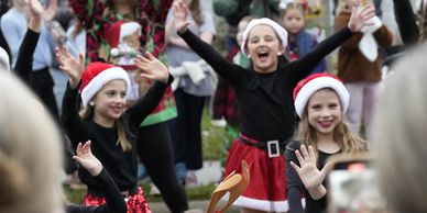 Children in festive costumes joyfully performing at a holiday event with an engaged audience.