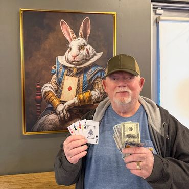 Man holding four aces and cash, standing before a framed white rabbit painting.
