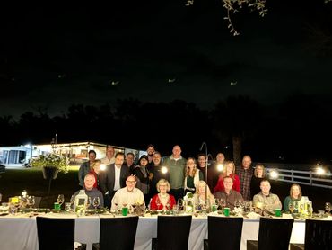 22 dinner guests sitting at the table for the Farm to Table dinner event, with market lights above.