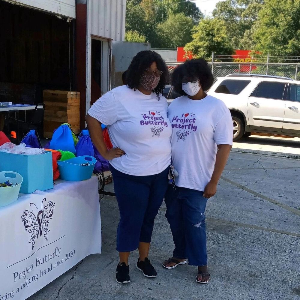Two women in "Project Butterfly" shirts stand by a table with colorful bags outdoors.