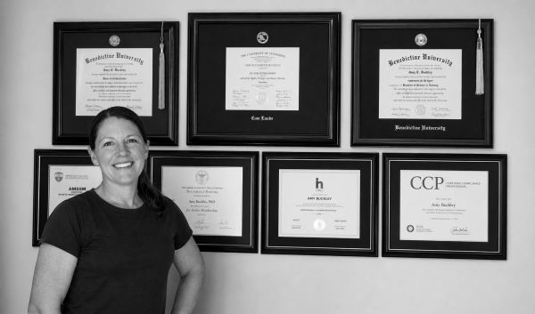 A woman stands smiling in front of framed diplomas and certificates on a wall.