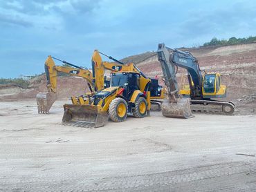 Three excavators parked on a construction site with a dirt hill in the background.