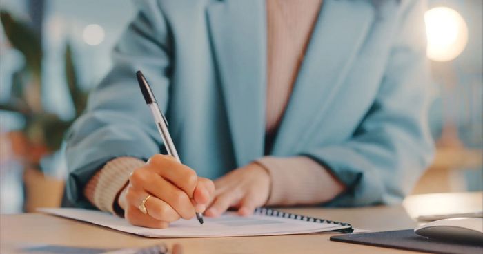 Person in a blue jacket writing on a spiral notebook with a pen.