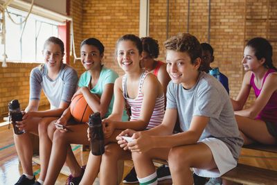 Teenagers sitting on a bench in a gym, holding water bottles and a basketball.