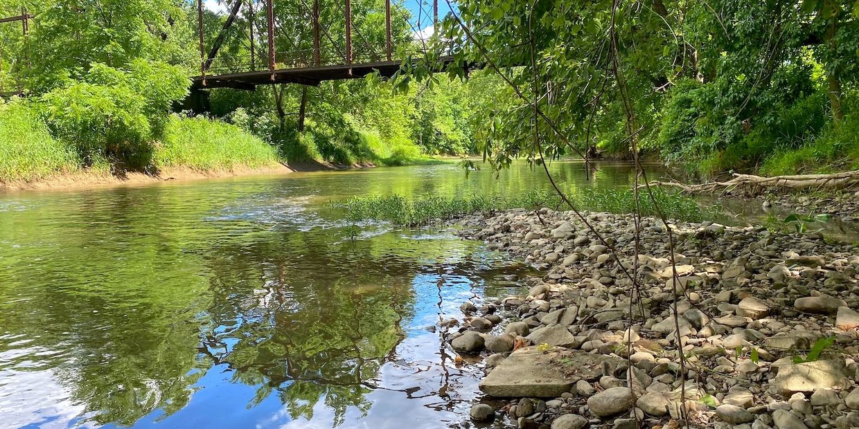 A peaceful river with a rocky shore and a rusty bridge overhead.